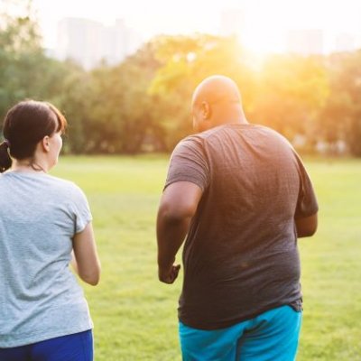 Two people, a man and woman, wear exercise gear outdoors in a green park and walk away from the camera.